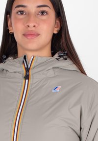 Giacca impermeabile grigio chiaro con cappuccio, dotata di una cerniera multicolore, patch del logo e un morbido rivestimento interno. Tessuto testurizzato.