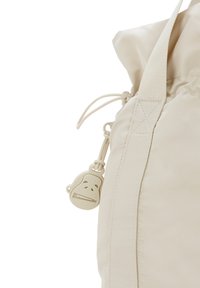 Beige rulle-toppväska gjord av ett slätt, lätt material. Har dragsko-stängning och ett litet dragkedjehandtag format som en apa.
