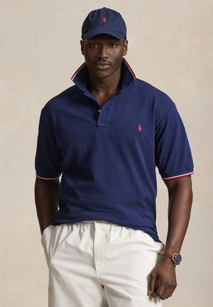 Mężczyzna ubrany w granatową koszulkę polo z czerwonym logo i obszyciem kołnierzyka, białe spodnie, granatową czapkę z czerwonym logo oraz brązowy zegarek na nadgarstku, stojący na neutralnym tle.
