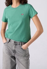 Femme portant un t-shirt vert Levi's et un jean gris délavé, les mains dans les poches, sur un fond uni.