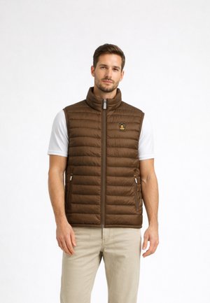 Uomo che indossa un gilet trapuntato marrone sopra una maglietta bianca a maniche corte e pantaloni beige, in piedi contro uno sfondo bianco semplice.