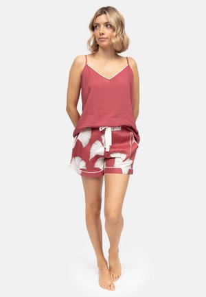 Mujer de pie descalza, vestida con un top sin mangas color rosa rosa y shorts a juego con estampado de hojas blancas y cordón, sobre un fondo liso.