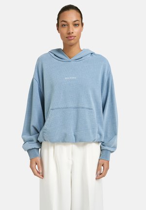 Woman wearing a loose light blue hoodie with "SOUL STUDIO" text and white wide-leg pants, standing against a plain background.