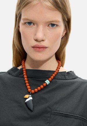 Młoda kobieta o jasnych włosach nosi czarny top i naszyjnik z dużych, okrągłych pomarańczowych koralików z zawieszką tukana w kolorach czarnym, białym i żółtym.