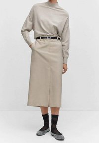 Beige wollen trui met hoge kraag, gecombineerd met een knielange, hoge taille rok met een split aan de voorkant en een zwarte riem. Zwarte doorzichtige kousen en flats.