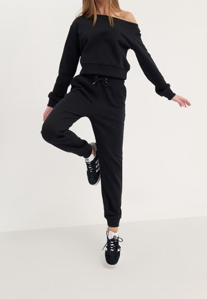 Frau balanciert auf einem Bein, trägt ein schwarzes schulterfreies Sweatshirt, schwarze Jogginghosen und schwarze Adidas-Sneakers mit weißen Streifen.