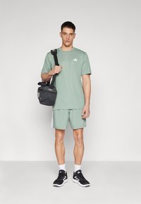 Grünes Sport-T-Shirt mit kleinem weißen Logo, passende grüne Shorts, schwarze Sneaker und eine schwarze Umhängetasche. Schlichtes Design, lässiger Look.