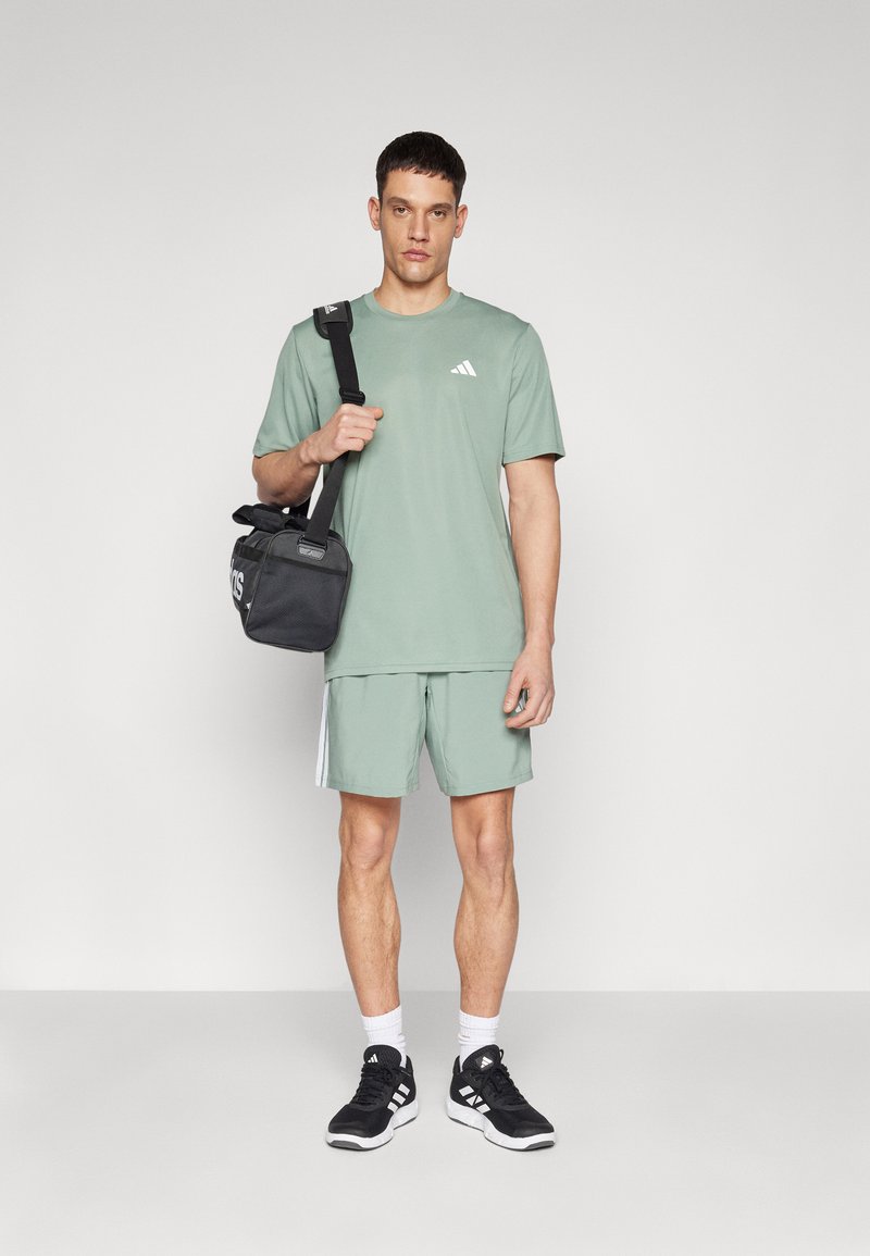 Camiseta atlética verde con un pequeño logotipo blanco, pantalones cortos verdes a juego, zapatillas negras y una bolsa de hombro negra. Diseño simple, aspecto casual.