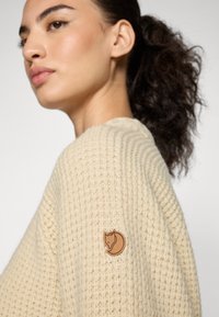 Chunky beige trui met een gestructureerde brei. Heeft een klein bruin dierenlogo op de linkerschouder. Eenvoudig, casual ontwerp.