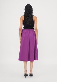 Falda morada hasta la rodilla con cintura elástica y lazos laterales, hecha de una tela suave. La modelo lleva una blusa negra de canalé y tacones.