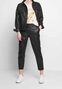 Chaqueta de cuero negra con dos bolsillos en el pecho, combinada con pantalones de cuero negros y una camiseta blanca con un estampado de cebra en amarillo y marrón. Zapatillas blancas.