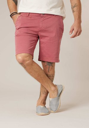 Man draagt vervaagde rode korte broek, lichtgrijze instapschoenen, wit shirt, armbanden en tatoeages op armen en benen, staand met gekruiste benen.