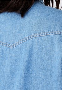 Light blue denim shirt featuring a pointed yoke and visible stitching. Fabric texture is soft with a smooth finish.
