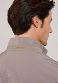 Giacca grigia chiara con colletto morbido e una fascia simile alla pelle scamosciata con il marchio "FACONNABLE" ricamato in beige. Setosa lucentezza del tessuto visibile.