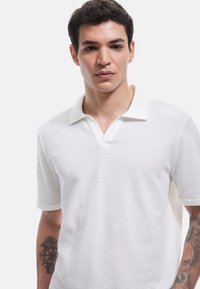 Hvid polo skjorte med et tekstureret design, featuring en V-hals og en kontrasterende ribkant. Korte ærmer med opsmøgte kanter.