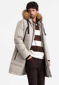 Cappotto beige imbottito con cappuccio foderato in pelliccia rimovibile, lacci neri e tasche con zip, indossato sopra una camicia a righe marroni e bianche.