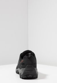 Black athletic shoe with a textured mesh upper, reinforced heel, and rugged rubber outsole. Features minimal red branding on the side.