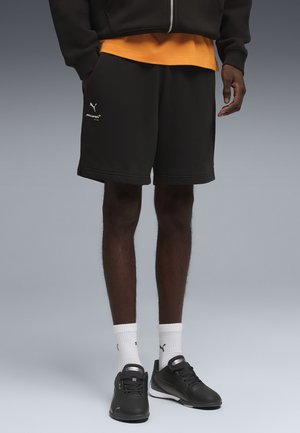 Persoon draagt zwarte Puma McLaren shorts, witte Puma sokken en zwarte Puma sneakers, staand tegen een neutrale grijze achtergrond.