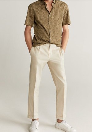 Man met een bruin, kort mouws overhemd met patroon, beige chino met omslag en witte sneakers, staand met handen in de zakken.