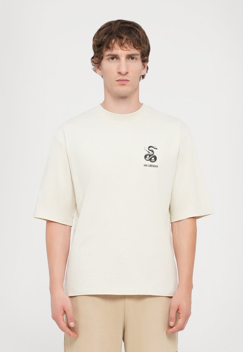 T-shirt en coton beige clair à manches courtes, avec un graphique de serpent noir sur le côté gauche de la poitrine. Design simple à col rond.