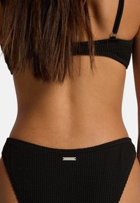 Top e slip bikini neri con trama strutturata, caratterizzati da un design a coste con un'etichetta argentata del marchio sulla fascia in vita, che mostrano linee pulite e uno stile classico.