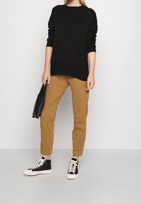 Personne portant un pull noir, un pantalon marron retroussé, des baskets noires et blanches, tenant une pochette noire, debout devant un fond blanc.