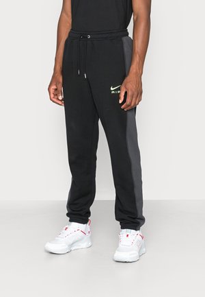 Homme portant un pantalon de jogging Nike Air noir et gris foncé avec une taille à cordon et des baskets blanches avec des accents rouges.