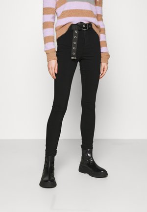 Tynne svarte jeans med en metallring-beltt, iført en myk, stripete genser i brun og lavendel. Støvler med tykk såle.