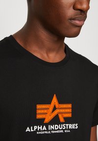Juodas medvilninis marškinėliai su tekstūruota oranžine logotipu su rodyklės dizainu ir prekės ženklu "ALPHA INDUSTRIES" baltu atspalviu, apvaliu kaklu.