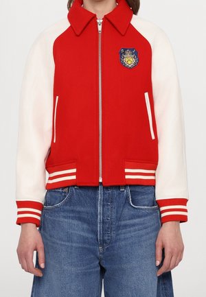 Persoon draagt een rood-wit varsityjack met gestreepte manchetten en zoom, geborduurd embleem op de borst, gecombineerd met een blauwe spijkerbroek.