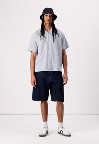 Cămașă cu mânecă scurtă, cu dungi în nuanțe de albastru deschis și alb, combinată cu pantaloni scurți din denim albastru închis. Pălărie bucket neagră și adidași asortați.