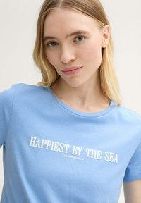 Camiseta de algodón azul claro con cuello redondo y mangas cortas. Texto "LOS MÁS FELICES JUNTO AL MAR" en blanco, con una textura suave.