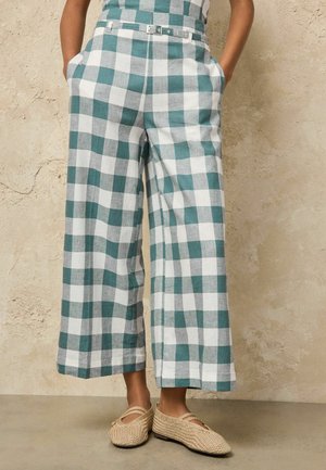 Person wearing green and white checkered wide-leg pants with a belt and beige woven flat shoes, standing against a textured wall.