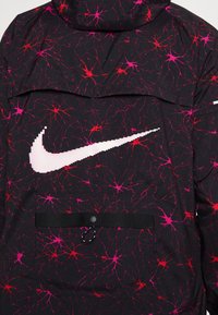 Zwarte jas met een levendig roze en rode abstracte print. Groot wit Nike-logo op de voorkant, met een zwarte zak en verstelbare banddetails.