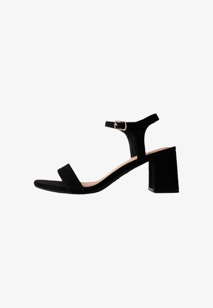 Czarna zamszowa sandałka na blokowanym obcasie, z odkrytymi palcami, paskiem wokół kostki i złotą klamrą, pokazana z profilu na białym tle.