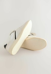 White sneakers with black accents, featuring a smooth leather upper, textured rubber outsole, and adjustable straps for closure.