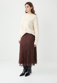 Pull en maille ivoire duveteux à manches longues associé à une jupe midi en dentelle marron. Des bottines noires à bout pointu complètent la tenue.