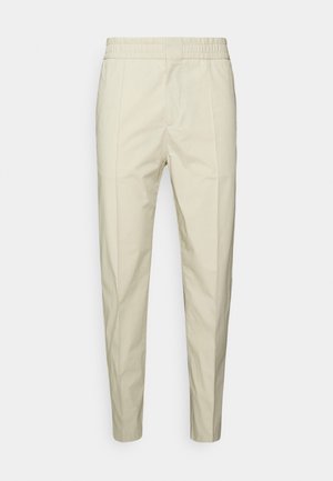 Beige katoenen broek met een elastische tailleband, taperende pijpen en twee voorkken. Soepele textuur zonder duidelijke patronen.