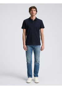 Marineblå polo t-shirt med krave, korte ærmer og stolpelukning. Kombination med lyseblå denim jeans og hvide sneakers. Neutral baggrund.