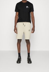 Muž v černém tričku DKNY, béžových kraťasech DKNY se stahovací šňůrkou, černých ponožkách a černých teniskách stojí na jednobarevném pozadí.