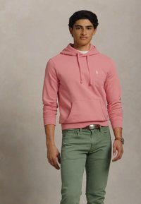 Νεαρός άντρας ντυμένος με ροζ φούτερ με λογότυπο και πράσινα παντελόνια, ουδέτερο φόντο.