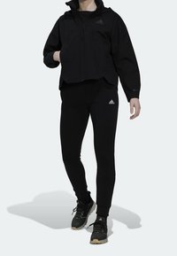 Personne vêtue d'une veste noire Adidas, de leggings et de baskets, se tenant avec une main dans la poche de la veste et un bras détendu.