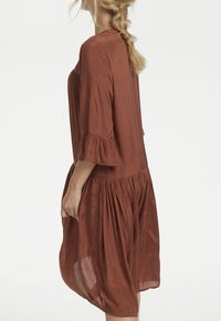 Brun, knælang kjole lavet af letvægtsstof. Har en afslappet pasform, 3/4 bell-ærmer og samlingdetaljer ved taljen.