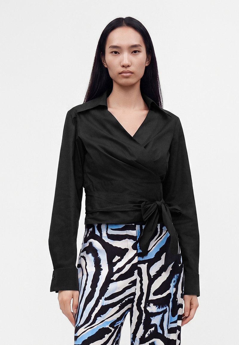 Femme aux longs cheveux noirs portant un chemisier cache-cœur noir avec un nœud attaché et un pantalon à motifs avec des rayures abstraites noires, blanches et bleues.