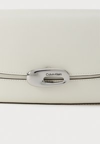 Calvin Klein hvid læder clutch med en glat tekstur, sølv oval lås og kontrasterende syninger langs kanten.