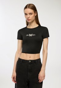 T-shirt nero cropped con maniche corte, caratterizzato da un design di una farfalla bianca e dal testo "Cose buone" sottostante. Texture liscia.