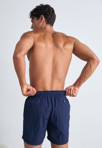 Jūros karinių šortų maudymosi, pagamintų iš lengvos medžiagos, su elastinga juosmens juosta ir užpakaline kišenė. Modelis be marškinėlių parodytas iš nugaros.