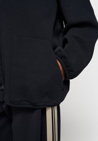 Schwarze Fleece-Jacke mit Knopfverschluss, einer großen Seitentasche und elastischen Bündchen. Strukturiertes Material mit sichtbaren Nahtdetails.