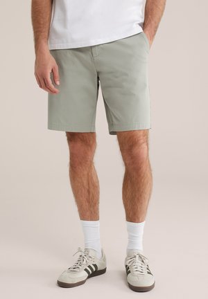 Muž nosi svetlošedé šortky, biele členkové ponožky a béžové tenisky s čiernymi pruhmi, stojí s jednou rukou v vrecku.