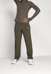 Olivengrønne joggere og en brun lynlåsjakke, begge lavet af glat materiale, med hvide logo accenter og en tætsiddende design.